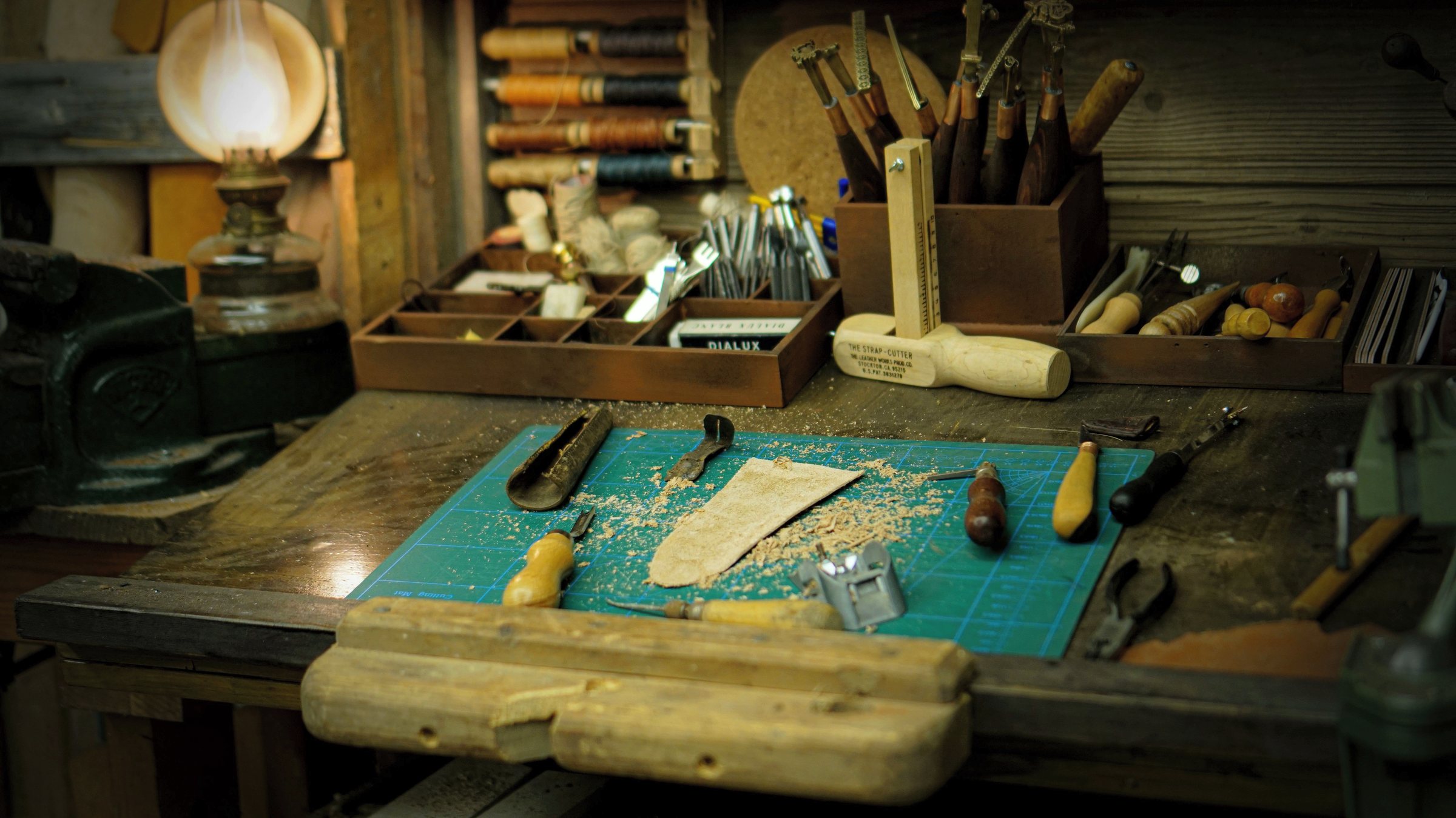 A craftsman's workbench at night, hand tools and a cutting mat lit by an oil lamp.