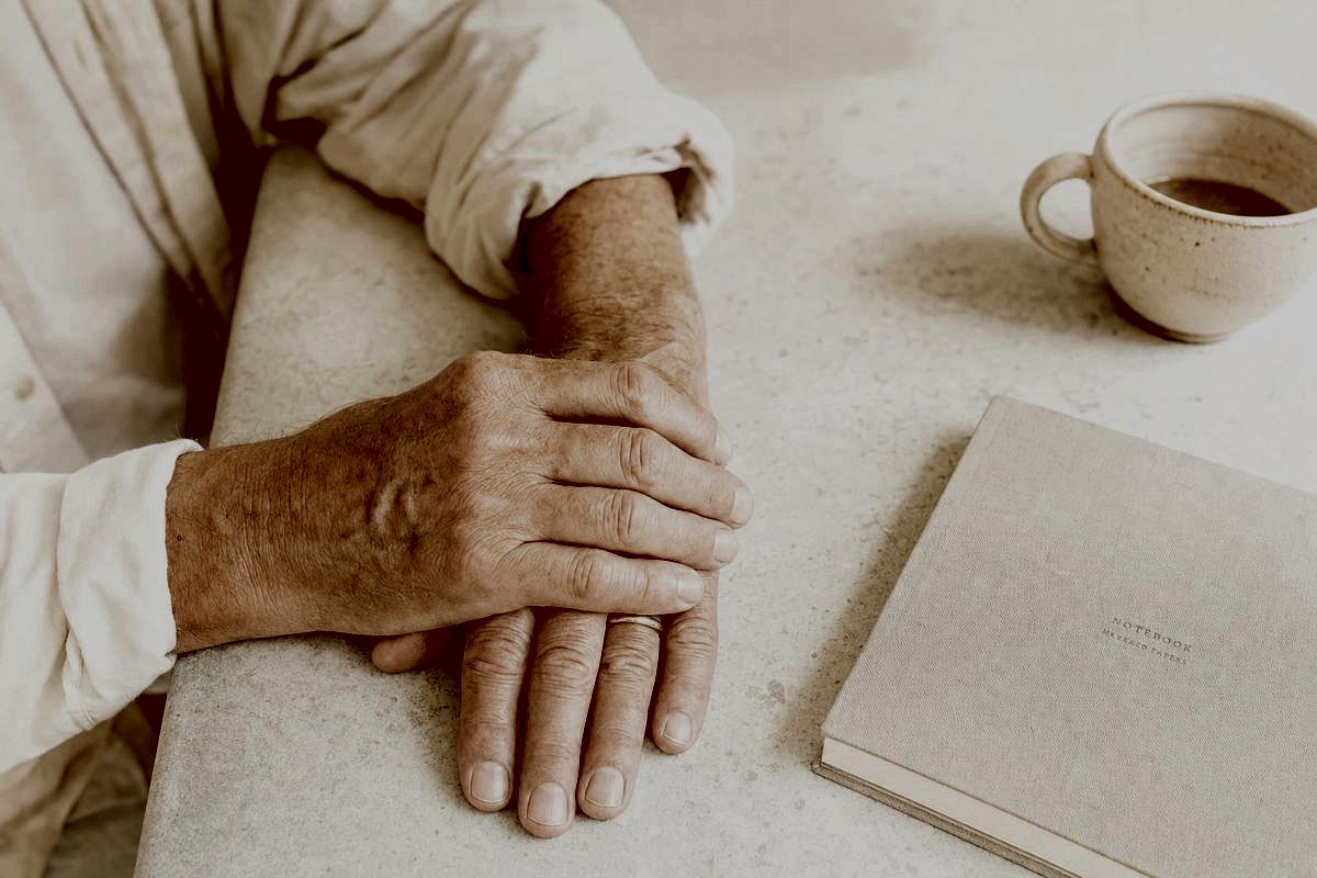 A pair of weathered hands resting still on a counter beside a closed notebook and a cup of coffee.